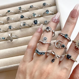 Puede incluir: Una colección de anillos de plata con piedras preciosas azules. Los anillos presentan varios diseños, incluyendo una luna creciente, una mariposa, un corazón y una corona. Los anillos se muestran en una caja de anillos de terciopelo blanco.