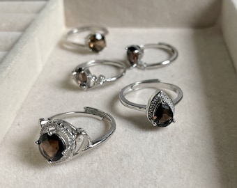 Puede incluir: Cinco anillos de plata con engastes de piedras preciosas de color marrón oscuro. Los anillos se muestran en una caja de joyería blanca.