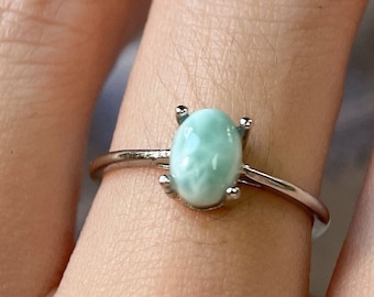 May include: A silver ring with a light blue oval-shaped gemstone. The ring is on a person's finger.