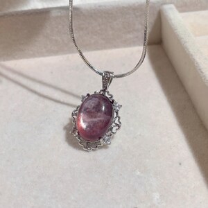 May include: A silver necklace featuring a pendant with a large, oval, pink gemstone. The gemstone is surrounded by a decorative silver frame with small, clear accent stones. The chain is a delicate silver snake chain.
