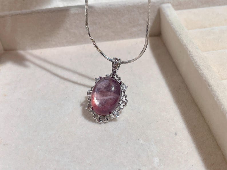 May include: A silver necklace featuring a pendant with a large, oval, pink gemstone. The gemstone is surrounded by a decorative silver frame with small, clear accent stones. The chain is a delicate silver snake chain.