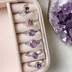 Peut inclure: Gros plan sur un présentoir à bagues avec six bagues serties d'améthystes. Les bagues sont disposées en rangée, la plus grande en haut et la plus petite en bas. Les bagues sont toutes en argent et ont des montures de styles différents. Les améthystes sont toutes de nuances de violet différentes.
