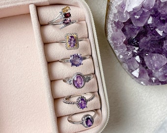 May include: A close-up of a ring display case with six rings featuring amethyst gemstones. The rings are arranged in a row, with the largest ring at the top and the smallest ring at the bottom. The rings are all silver and have different styles of settings. The amethyst gemstones are all different shades of purple.