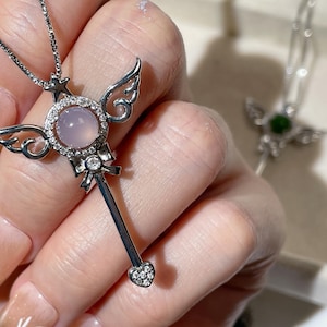 May include: A silver necklace featuring a pink gemstone pendant shaped like a wand with angel wings. The pendant is decorated with small clear stones.