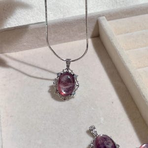 May include: A silver-toned necklace featuring an oval-shaped, translucent pink gemstone pendant. The pendant is set in a decorative silver-toned metal frame with small clear accent stones. A thin, smooth silver-toned chain is attached to the pendant.
