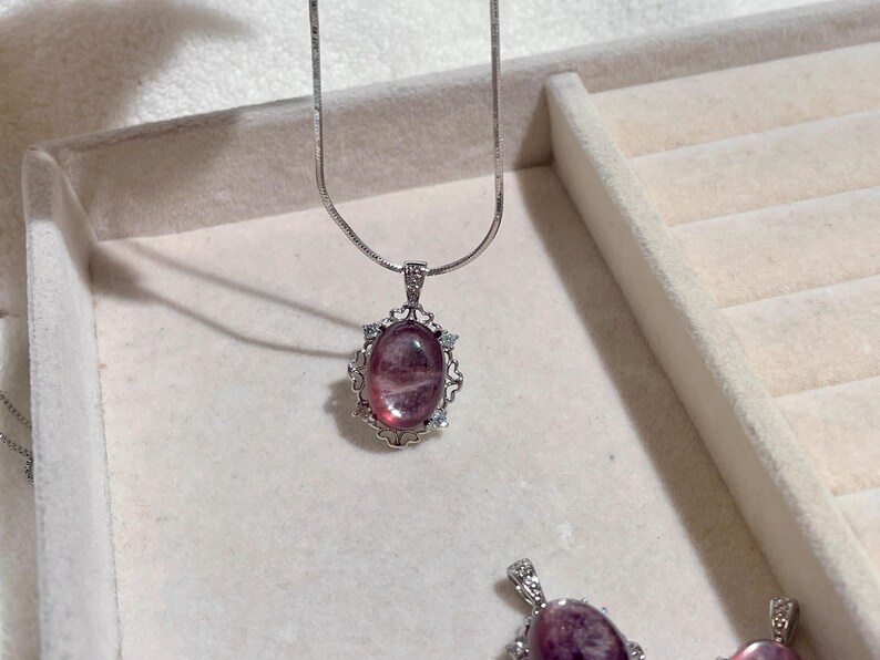 May include: A silver-toned necklace featuring an oval-shaped, translucent pink gemstone pendant. The pendant is set in a decorative silver-toned metal frame with small clear accent stones. A thin, smooth silver-toned chain is attached to the pendant.