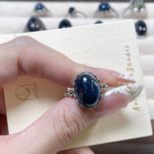 May include: A silver-toned ring featuring a large, oval-shaped, dark blue cabochon gemstone with subtle internal marbling. The setting is accented with small, clear, round-cut stones along the band, which is displayed on a cream-colored card.