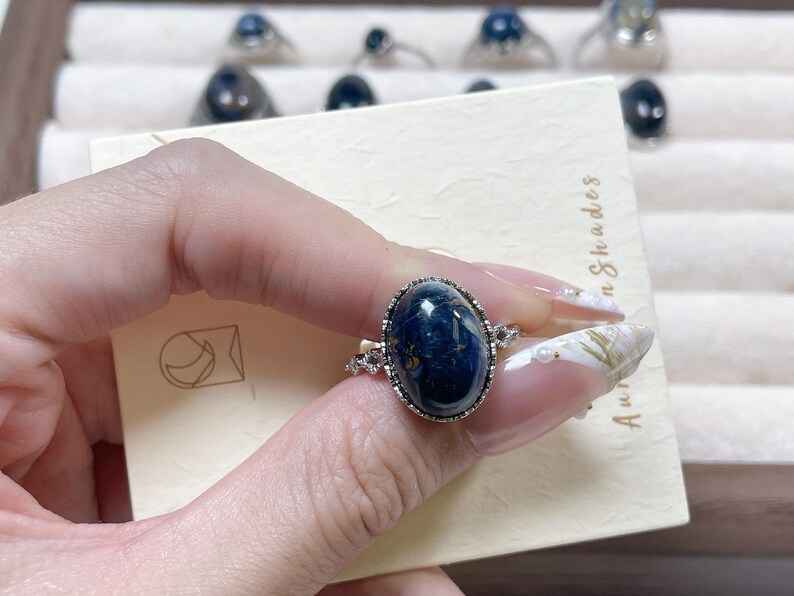 May include: A silver-toned ring featuring a large, oval-shaped, dark blue cabochon gemstone with subtle internal marbling. The setting is accented with small, clear, round-cut stones along the band, which is displayed on a cream-colored card.