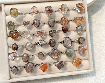 May include: A collection of silver rings with various shapes and sizes of clear and brown crystal stones. The rings are displayed in a white box with velvet lining.