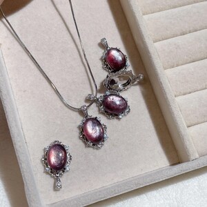May include: A jewelry set featuring a necklace, earrings, and a ring. Each piece has an oval, purplish-pink gemstone with a silver-colored, ornate setting. The jewelry is displayed in a light-colored, velvet-lined jewelry box.