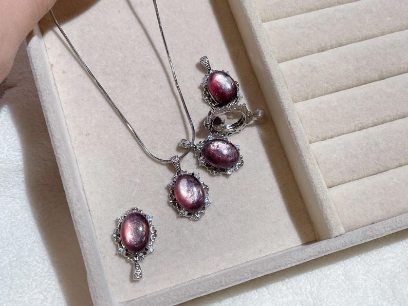 May include: A jewelry set featuring a necklace, earrings, and a ring. Each piece has an oval, purplish-pink gemstone with a silver-colored, ornate setting. The jewelry is displayed in a light-colored, velvet-lined jewelry box.