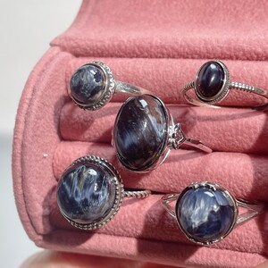 May include: Five silver rings featuring polished, oval and round cabochon gemstones with swirling blue, gray, and dark brown patterns. The rings have varied band designs, including twisted and beaded textures, displayed in a pink velvet jewelry box.
