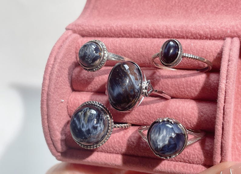 May include: Five silver rings featuring polished, oval and round cabochon gemstones with swirling blue, gray, and dark brown patterns. The rings have varied band designs, including twisted and beaded textures, displayed in a pink velvet jewelry box.