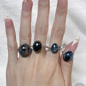 May include: Four silver-toned rings featuring oval-shaped, dark blue and black polished gemstones. Each ring has a unique metal band design, including intricate filigree and smooth settings, worn on four fingers of a single hand.