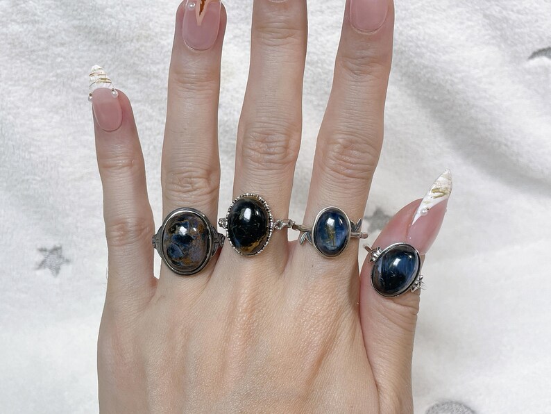 May include: Four silver-toned rings featuring oval-shaped, dark blue and black polished gemstones. Each ring has a unique metal band design, including intricate filigree and smooth settings, worn on four fingers of a single hand.