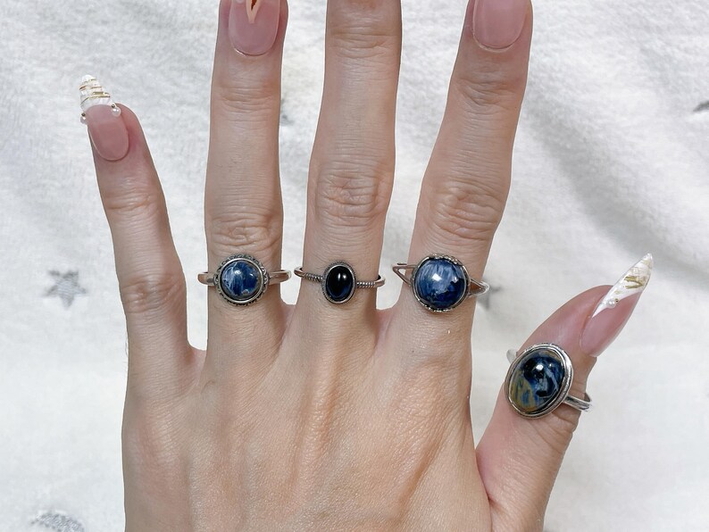 May include: Four silver-toned rings worn on fingers. Three rings feature round or oval cabochon stones with blue and black marbled patterns. One ring in the center features a smaller, smooth, solid black oval stone set in a thin band.