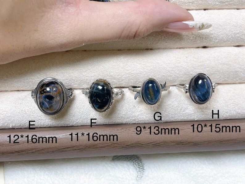 May include: Four silver-toned rings featuring oval blue and black gemstones, labeled E, F, G, and H. Ring E is 12x16mm, F is 11x16mm, G is 9x13mm, and H is 10x15mm. Each ring has a unique metal band design holding the polished cabochon stone.