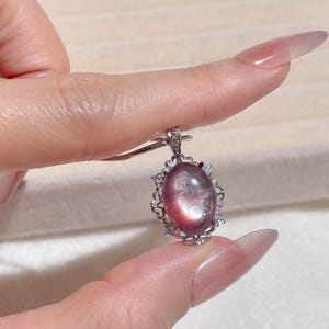 May include: A silver pendant necklace featuring an oval, pinkish-purple gemstone. The gemstone is set in a decorative silver frame with small, clear accent stones. The necklace is held up by a silver chain.