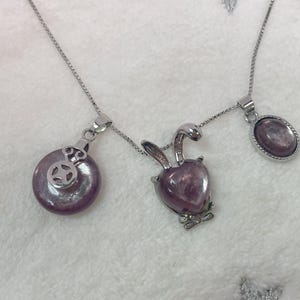 Puede incluir: Un collar de cadena de plata con tres colgantes. Los colgantes est&aacute;n hechos de piedra p&uacute;rpura y tienen diferentes formas, incluyendo un c&iacute;rculo, un coraz&oacute;n y un &oacute;valo.