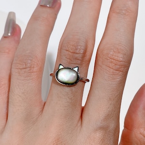 Könnte beinhalten: Ein silberner Ring mit einem weißen, katzenförmigen Muschelkamee. Der Ring befindet sich an einem Finger.