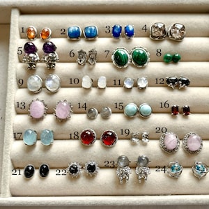 Puede incluir: Una caja blanca que contiene 24 pares de pendientes con diferentes piedras preciosas y diseños. Los pendientes están dispuestos en filas en una caja blanca con los números del 1 al 24 etiquetando cada par.