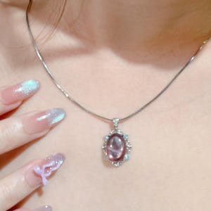 May include: A silver-toned necklace featuring a delicate chain and an oval-shaped pendant. The pendant contains a smooth, translucent purple stone set within a decorative silver frame accented with small, clear, sparkling stones.