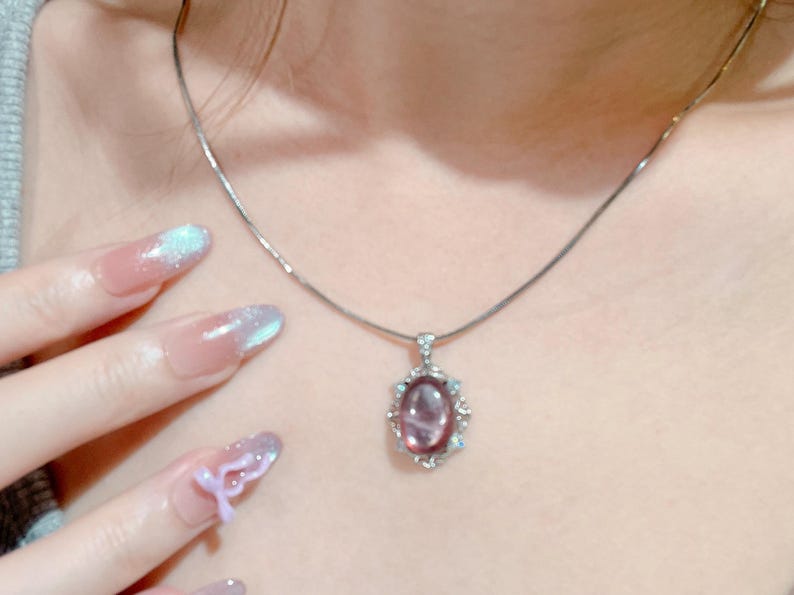 May include: A silver-toned necklace featuring a delicate chain and an oval-shaped pendant. The pendant contains a smooth, translucent purple stone set within a decorative silver frame accented with small, clear, sparkling stones.