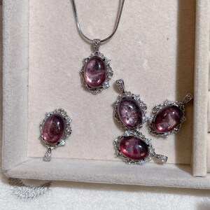 May include: A collection of jewelry pieces featuring oval-shaped, reddish-purple gemstones. The set includes a pendant necklace and several matching pendants, all set in ornate, silver-toned frames. The gemstones have a translucent quality, and the jewelry is displayed on a light-colored surface.