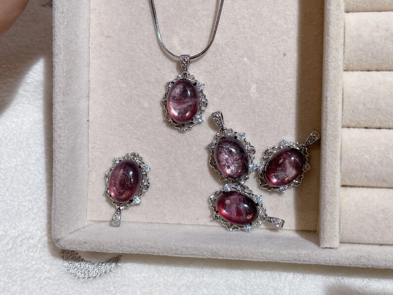 May include: A collection of jewelry pieces featuring oval-shaped, reddish-purple gemstones. The set includes a pendant necklace and several matching pendants, all set in ornate, silver-toned frames. The gemstones have a translucent quality, and the jewelry is displayed on a light-colored surface.