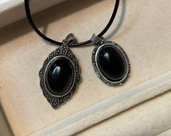 Puede incluir: Un collar de cordón negro con dos colgantes de plata con piedras ovaladas negras. Los colgantes tienen intrincados diseños de plata.