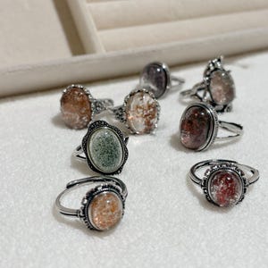 Könnte beinhalten: Eine Sammlung von Edelsteinringen mit silberfarbenen Bändern. Die Ringe zeigen ovale Steine in Orange-, Grün- und Rottönen. Die Ringe werden auf einer weißen Oberfläche präsentiert, um ihre detaillierten Designs und Farben hervorzuheben.