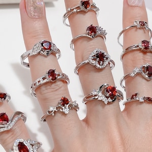 Op de afbeelding: Een handmodel draagt ​​een verscheidenheid aan zilveren ringen met rode edelsteenaccenten. De ringen hebben verschillende stijlen, waaronder hart-, bloemen- en geometrische ontwerpen.