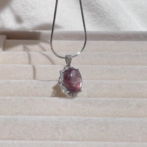 May include: A silver-toned necklace featuring a smooth, oval-shaped purple gemstone pendant. The stone is set in a decorative silver-toned frame accented with small, clear, sparkling stones along the edges and the bail.