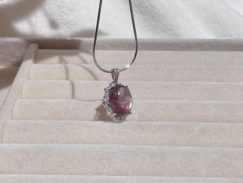May include: A silver-toned necklace featuring a smooth, oval-shaped purple gemstone pendant. The stone is set in a decorative silver-toned frame accented with small, clear, sparkling stones along the edges and the bail.