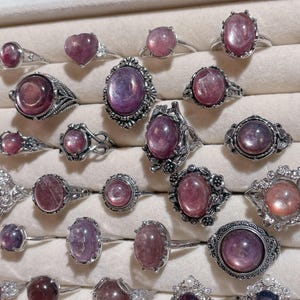 May include: A collection of silver rings with various pink and purple gemstones. The rings feature different designs, including heart-shaped stones and ornate settings. The gemstones have a translucent appearance, and the rings are displayed in a white jewelry tray.