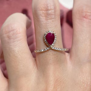 Könnte beinhalten: Ein goldener Ring mit einem tropfenförmigen Rubin, der von kleineren Diamanten umgeben ist. Der Ring hat eine zarte, gebogene Band mit Diamanten, die entlang der Kanten besetzt sind.