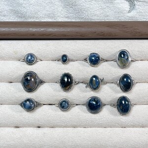 May include: A collection of twelve silver rings featuring oval and round blue tiger's eye gemstones. The rings are arranged in three rows, showcasing various metal band designs, including smooth, beaded, and filigree settings with polished stones.