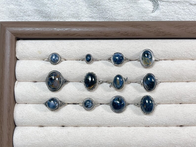 May include: A collection of twelve silver rings featuring oval and round blue tiger's eye gemstones. The rings are arranged in three rows, showcasing various metal band designs, including smooth, beaded, and filigree settings with polished stones.
