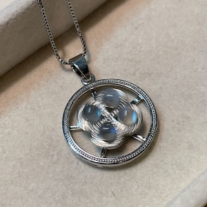 Op de afbeelding: Een zilveren ketting met een ronde hanger met een wit maanstenen ontwerp. De hanger heeft een delicaat, ingewikkeld patroon en is omgeven door een zilveren rand.