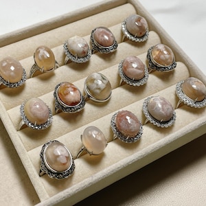 May include: A collection of 13 rings featuring oval-shaped agate stones set in silver. The stones display a mix of white, pink, and brown colours. The rings are presented in a white velvet-lined box.