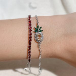 May include: Two bracelets on a wrist. One is a silver tennis bracelet with red gemstones. The other is a silver bracelet with a pineapple charm. The pineapple charm has a yellow centre stone and green leaves.