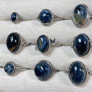 May include: A collection of nine silver rings featuring oval and round blue tiger eye gemstones. Each ring displays a unique chatoyant pattern with deep blue and golden-brown streaks, set in polished silver bands with various decorative metal settings.
