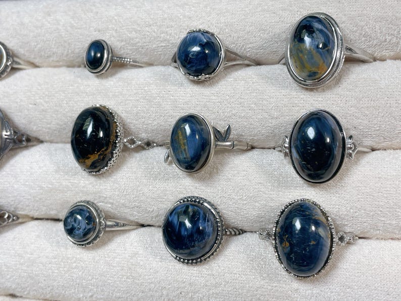 May include: A collection of nine silver rings featuring oval and round blue tiger eye gemstones. Each ring displays a unique chatoyant pattern with deep blue and golden-brown streaks, set in polished silver bands with various decorative metal settings.