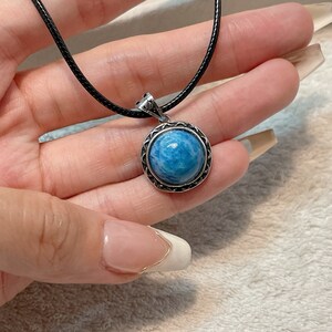 May include: A round, blue pendant necklace with a silver-colored bezel and a black cord. The pendant has a textured, light blue stone. The necklace is held in a hand.
