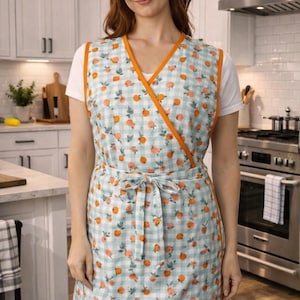 May include: A light blue and white checkered apron with orange trim and orange fruit designs. The apron is tied at the waist with a matching fabric tie. The apron is worn over a white shirt.