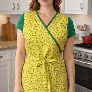 May include: A yellow apron with a green trim and a pattern of green bean-like shapes. The apron is tied at the waist with a matching fabric tie. The model is wearing a green t-shirt underneath the apron. The background is a kitchen.