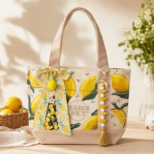 May include: A beige canvas tote bag featuring a yellow lemon print and the text Trader Joe's. The bag is decorated with a patterned fabric bow, a small yellow lemon charm, and a vertical strand of yellow wooden beads with a tassel at the bottom.