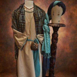 May include: A tan-colored costume with a patterned shawl, teal sash, and beaded necklace. A matching head covering and scarf rest on a carved wooden stand. The costume is displayed on a mannequin.