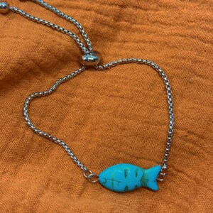 May include: An adjustable silver bracelet featuring a turquoise fish charm with green detailing. The charm is attached to a fine chain. The bracelet is shown against an orange fabric backdrop.