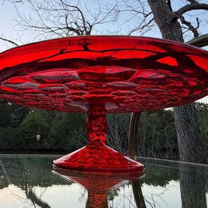Puede incluir: Un vibrante soporte para pasteles de vidrio rojo con un patrón texturizado similar a burbujas en la parte inferior del plato. El soporte tiene una parte superior ancha y plana y una base de pedestal. El soporte se refleja en una superficie de vidrio.